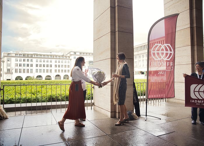 Twee vrouwen begroeten elkaar buiten in de zuilengang, naast de congresbanner aan de ingang