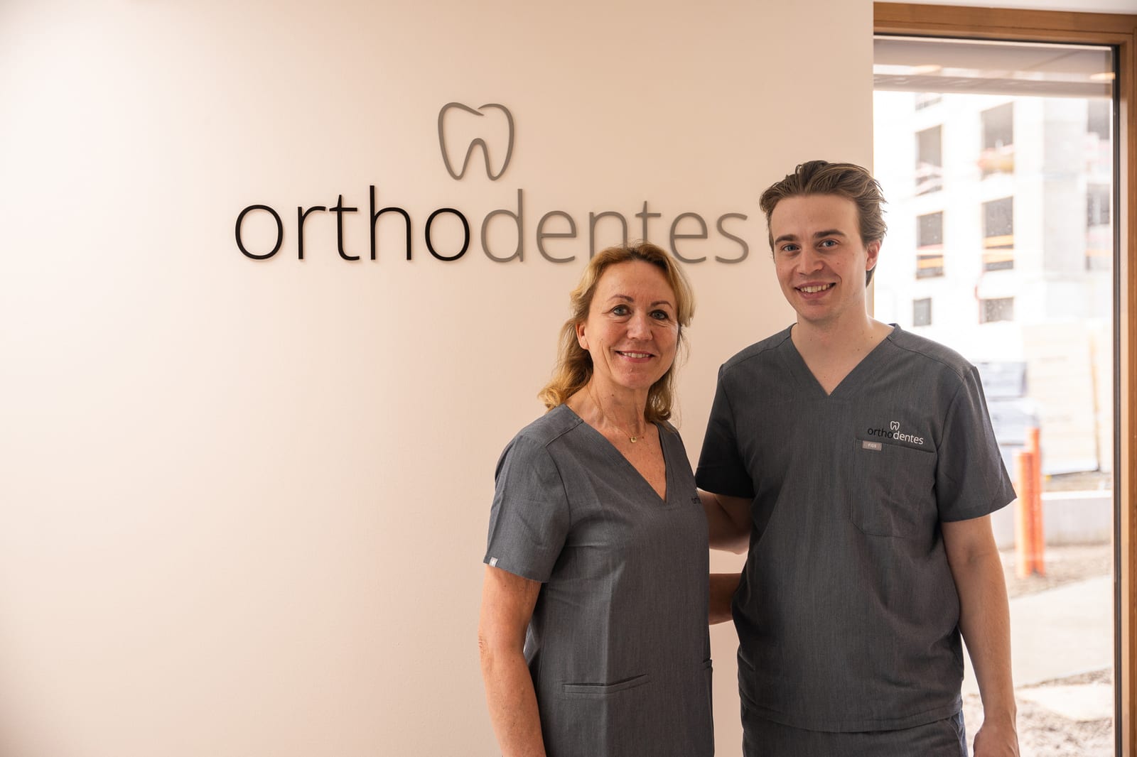 Twee behandelaars van Orthodentes bij het logo aan de muur van hun praktijk